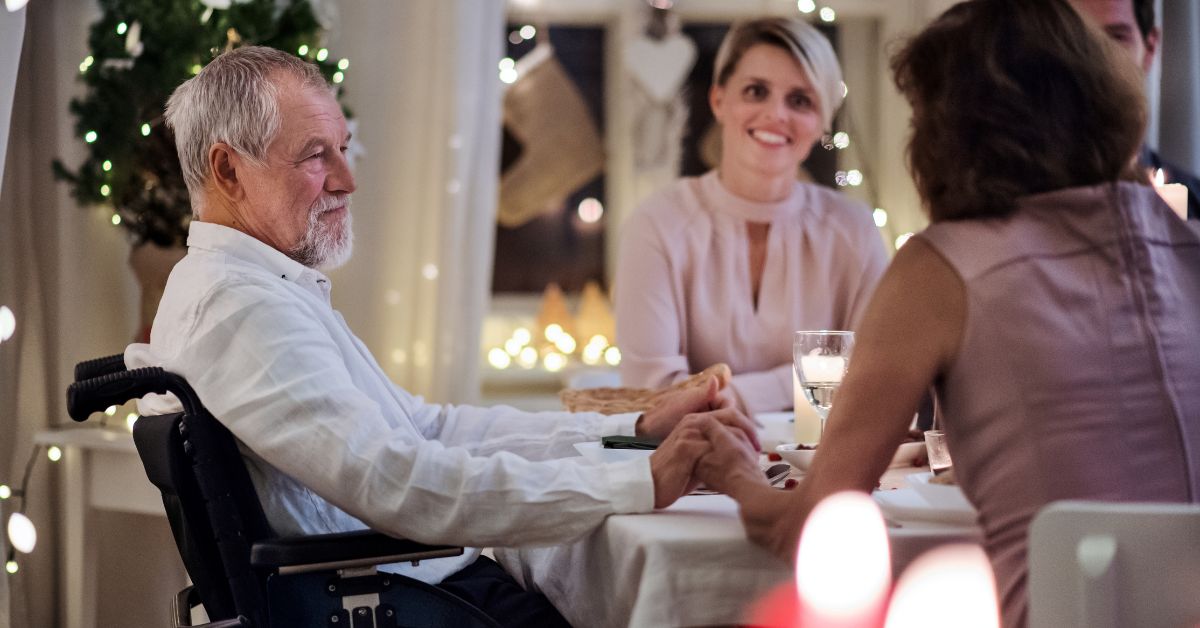 An elderly man in a wheelchair holds hands with a family member across a festive dining table, surrounded by smiling loved ones and warm string lights.