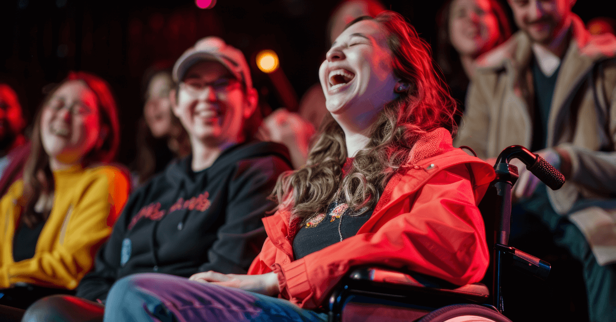 A woman in a wheelchair laughs with others in a seated audience at an indoor event, with colorful stage lighting.