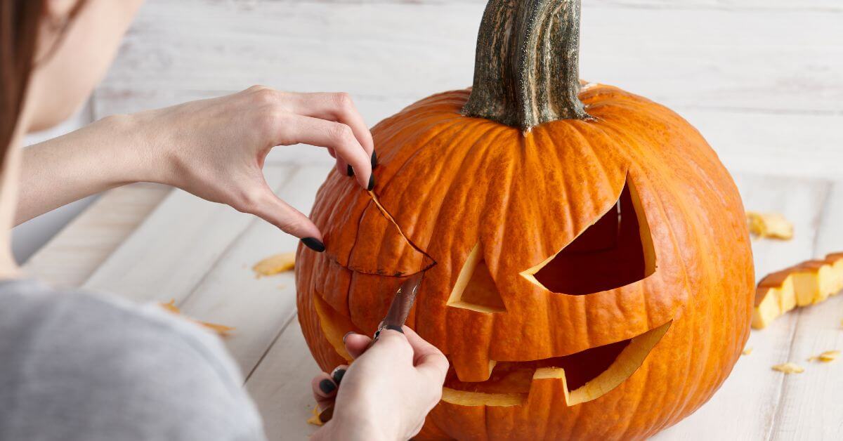 A person carves a face into an orange pumpkin, removing a triangular piece to create a Jack-o-lantern on a light-colored surface.
