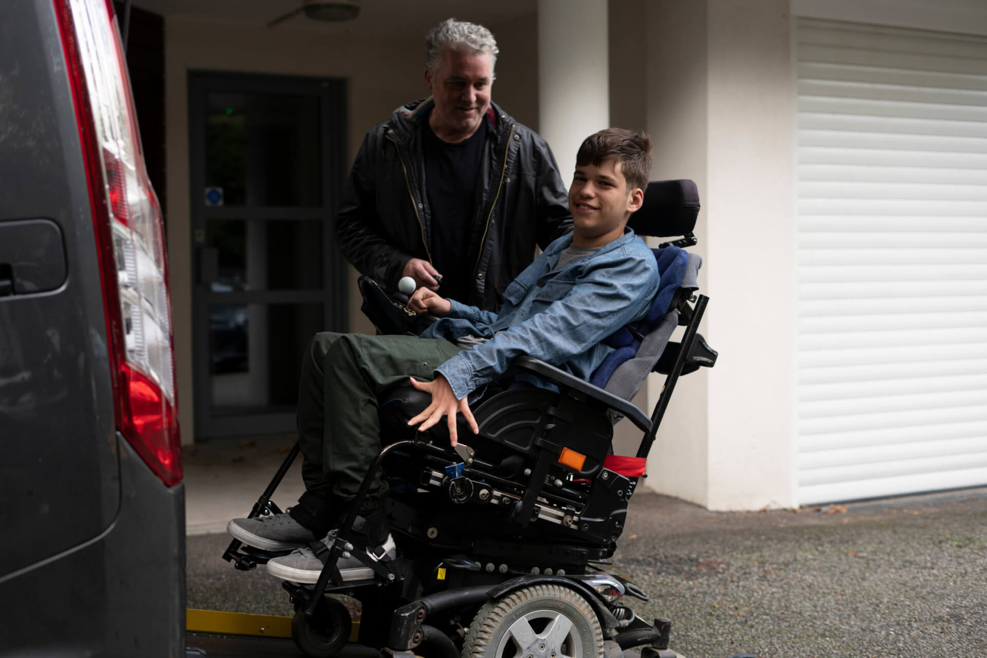 A young person in a motorized wheelchair uses a lift to board a van, assisted by an adult standing behind them outside a building—demonstrating the accessibility and convenience offered through the Buy Direct™ Program.