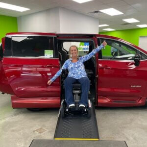 A woman sits in a wheelchair lift seat extended from a red wheelchair van inside a building, demonstrating the vehicle’s accessibility features.