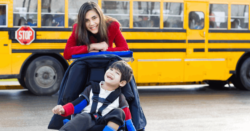 A woman stands behind a smiling boy in a wheelchair, with a yellow school bus in the background.