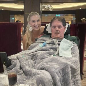 A woman stands and smiles behind a man in a wheelchair, covered with a gray blanket and attached to a ventilator, as they enjoy time together in a restaurant after arriving in their wheelchair van.