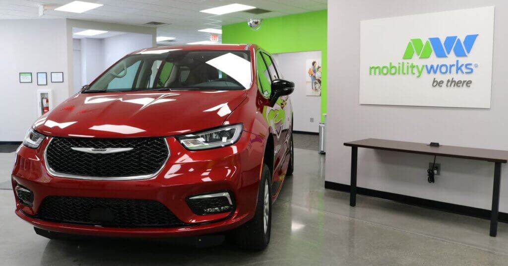 A red Chrysler minivan is parked inside a MobilityWorks showroom with a white and green MobilityWorks sign on the wall.