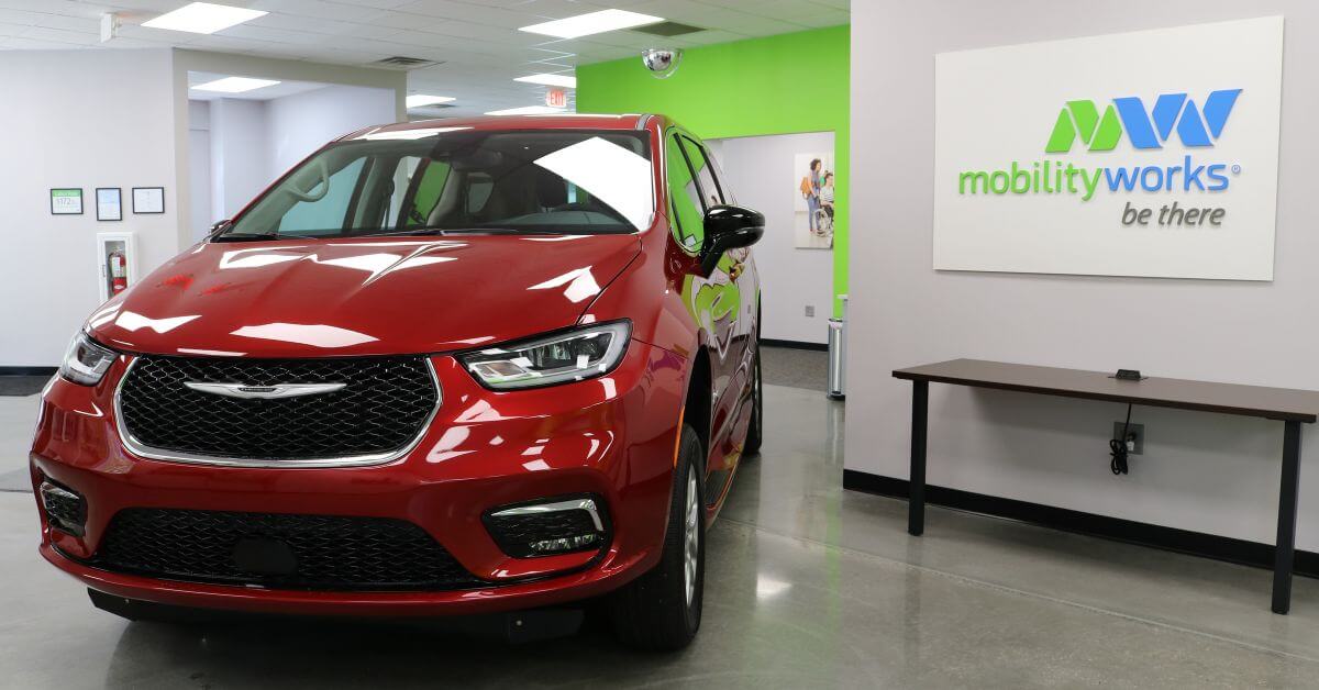 A red Chrysler minivan is parked inside a MobilityWorks showroom with a white and green MobilityWorks sign on the wall.