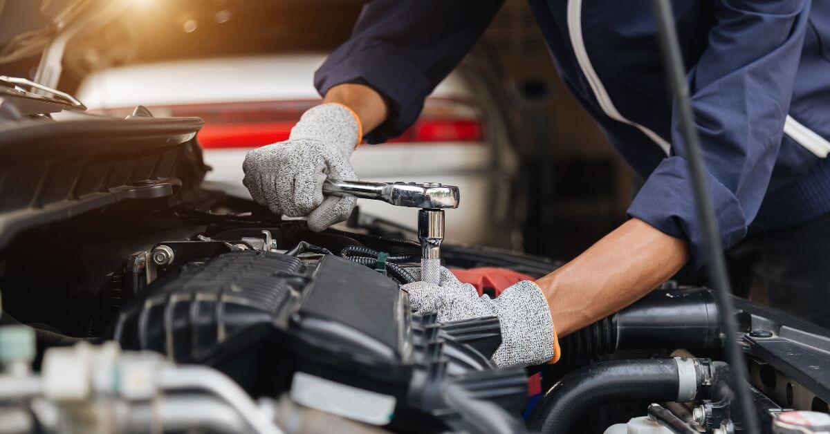 Person wearing gloves uses a ratchet wrench to work on a wheelchair accessible car engine in a brightly lit garage.