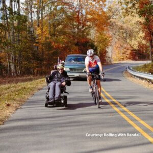 Two people travel on a winding road: one in a motorized wheelchair with a small flag, the other on a bicycle. Autumn trees and a car, possibly a wheelchair van, are in the background.
