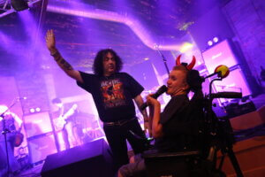 A man in an AC/DC shirt raises his arm on stage while another man in a wheelchair, wearing red devil horns, sings into a microphone—celebrating Rare Disease Day as the band rocks out in the background.