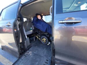 A person in a wheelchair sits inside a van with a ramp, covered by a dark blanket. The van door is open, revealing the accessible interior—highlighting mobility challenges often faced on Rare Disease Day.