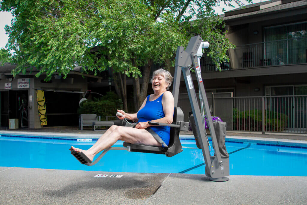 Una mujer mayor con un traje de baño azul está sentada en un elevador de piscina, mientras la bajan a una piscina al aire libre en un complejo residencial, sonriendo y sujetando una cuerda.
