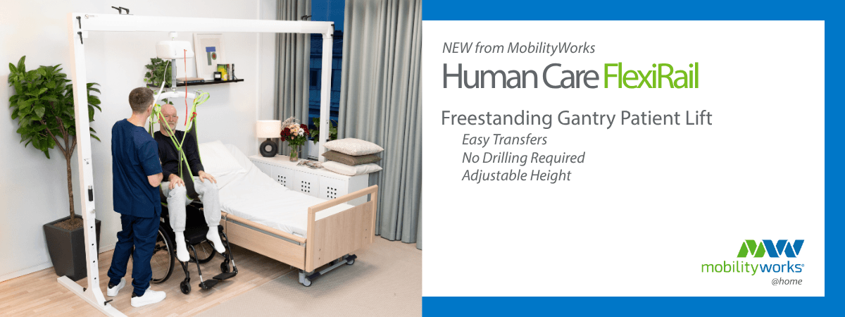 A caregiver assists a seated man using a gantry patient lift next to a hospital bed in a well-lit room with plants and shelves.