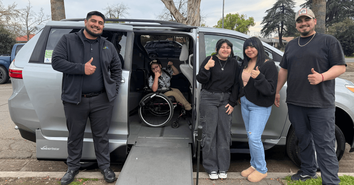Five people pose next to a van with a wheelchair ramp; one person in a wheelchair is inside the van, and the others stand outside, all giving a thumbs-up gesture.