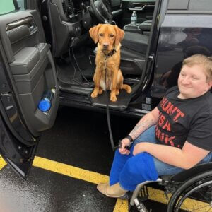A person in a wheelchair holds a leash attached to a dog sitting on the open door frame of a parked wheelchair van.