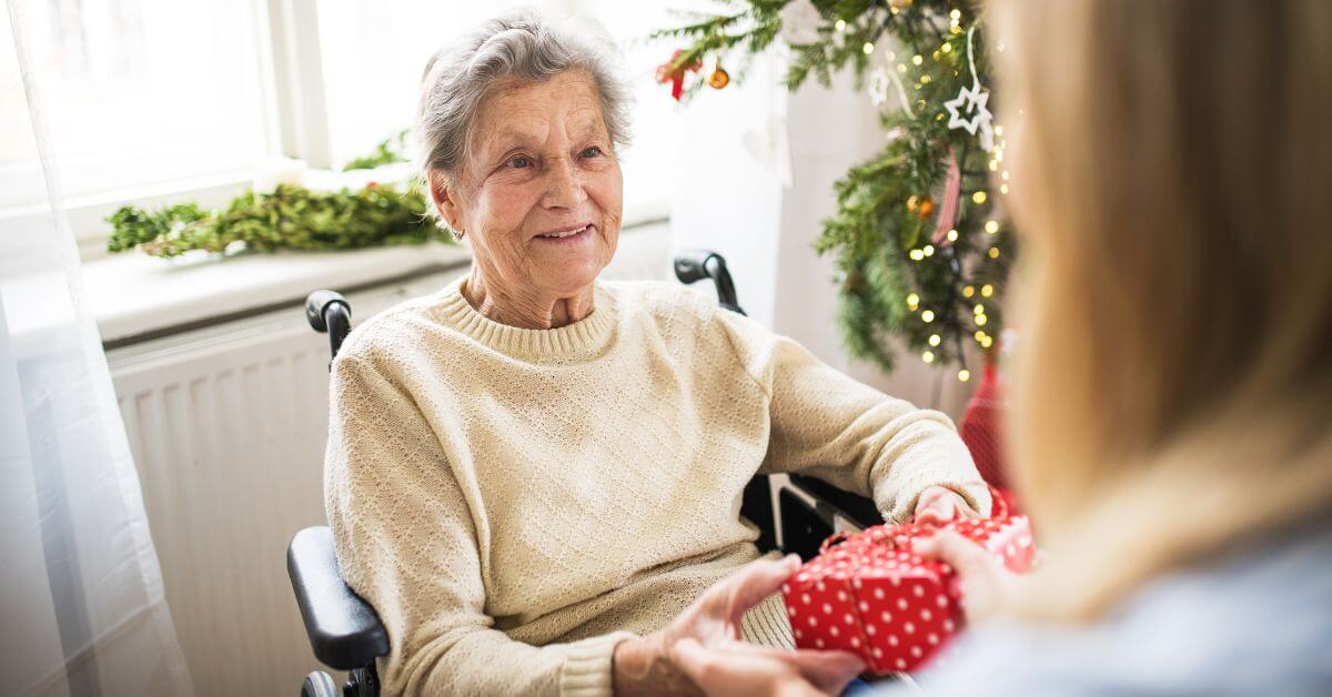 An elderly woman in a wheelchair receives a red polka dot gift from another person beside a decorated Christmas tree.