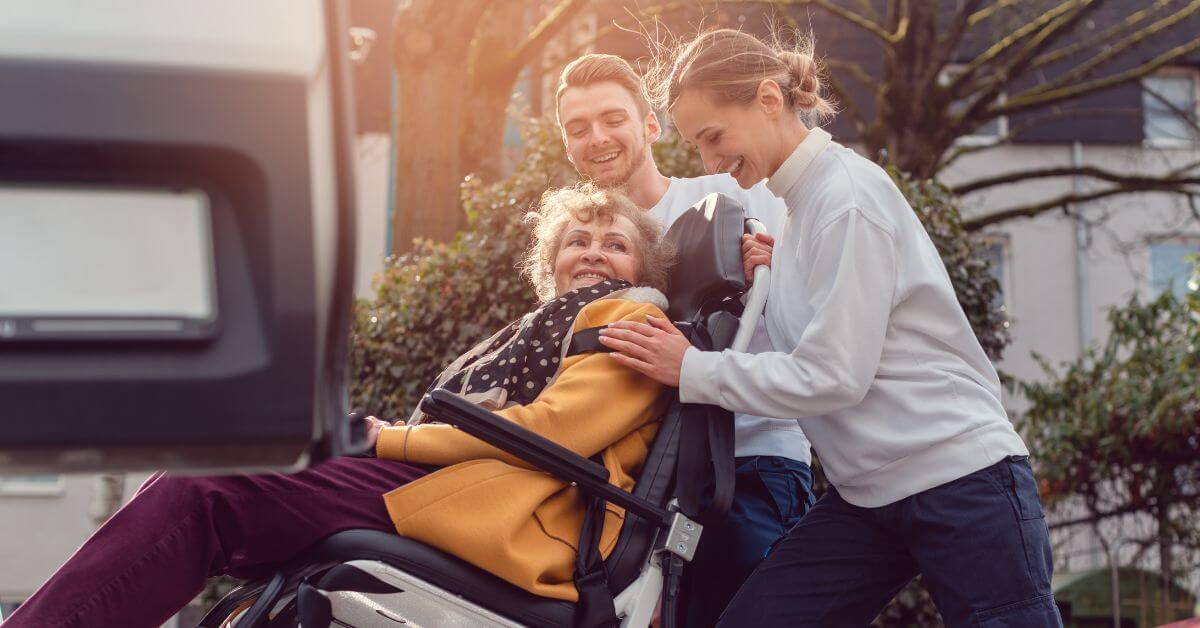 An elderly woman in a wheelchair is being assisted outdoors by two smiling younger adults, possibly volunteers from disability nonprofits. Trees and buildings are visible in the background.
