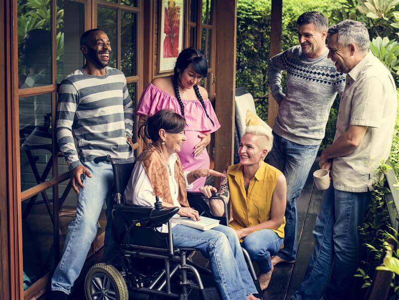 Six adults, including a woman in a wheelchair, gather on a porch, chatting and smiling in a relaxed, outdoor setting with greenery in the background.