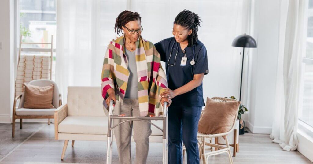 A young Black caregiver in blue scrubs gently assists an elderly Black woman using a walker in a bright, modern living room.