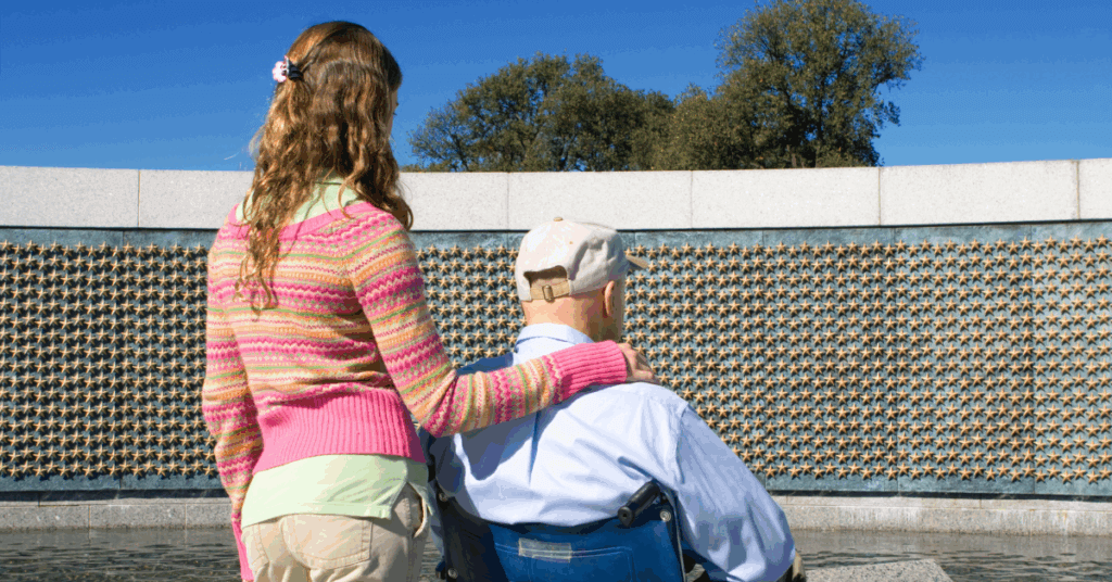 A young woman stands beside an older man in a wheelchair at a memorial wall decorated with gold stars, under a clear blue sky.