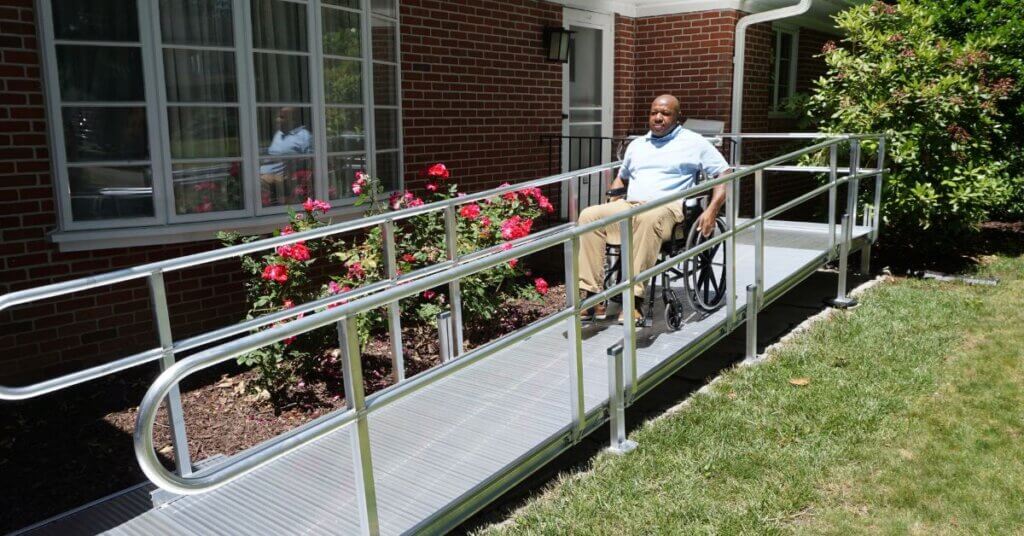 A person in a wheelchair uses a metal accessibility ramp outside a brick building with flowers and bushes nearby.