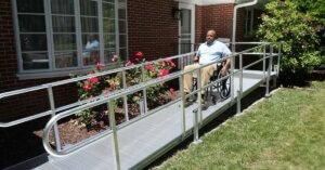 A person in a wheelchair uses a metal accessibility ramp outside a brick building with flowers and bushes nearby.