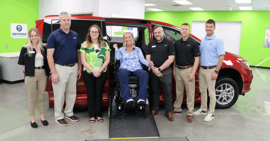 Seven people stand in front of a red accessible van; one woman sits in a power wheelchair on a ramp extending from the van’s side.