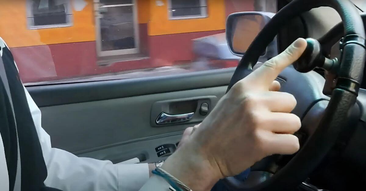 A disabled driver uses a steering wheel knob on the steering wheel while driving, with a view of a building through the driver’s side window.