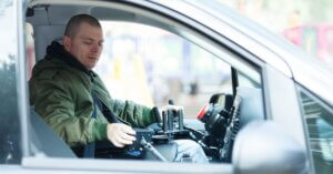 A man in a green jacket sits in the driver's seat of a car, operating specialized adaptive driving controls with both hands. He looks down at the controls with a focused expression.