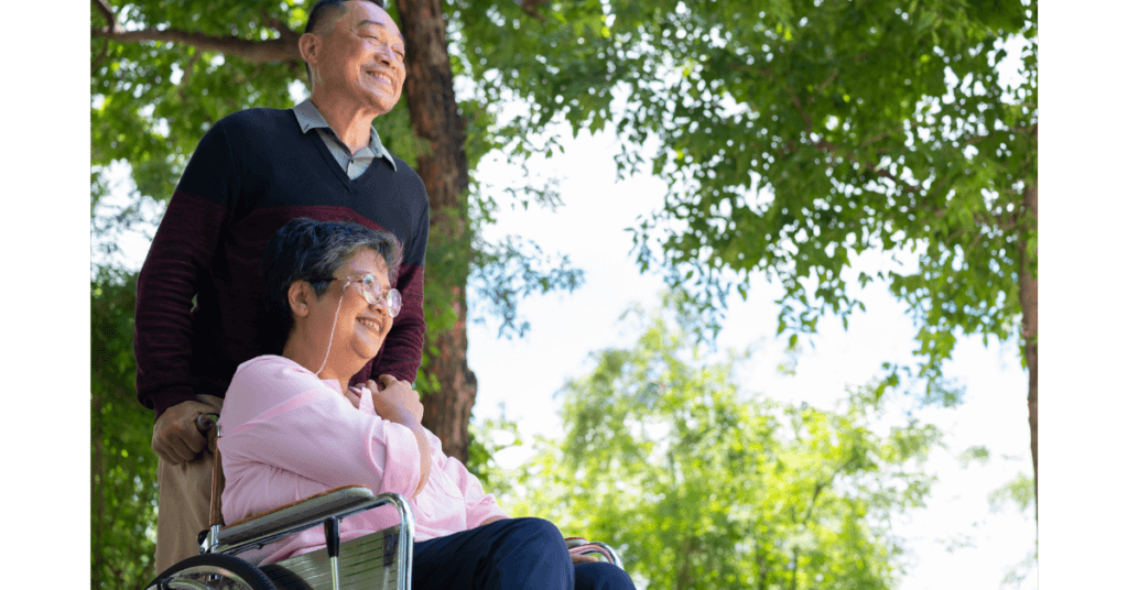 An older man stands behind an older woman in a wheelchair, both smiling outdoors with green trees in the background.