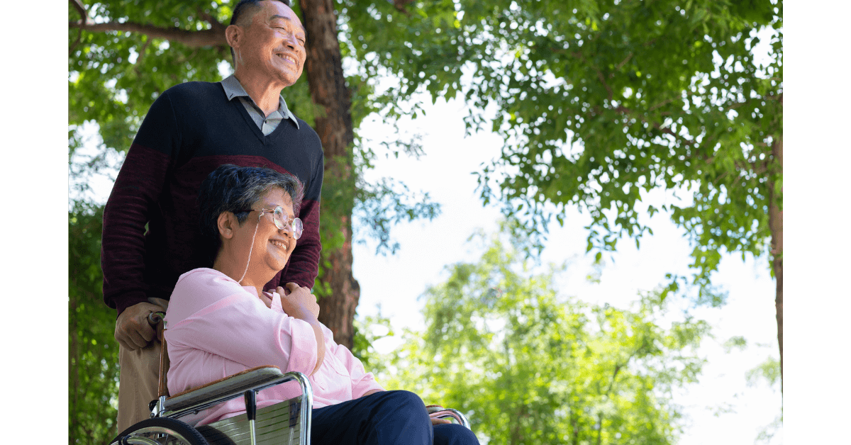 An older man stands behind an older woman in a wheelchair, both smiling outdoors with green trees in the background.