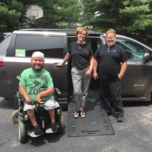 Three adults pose by a wheelchair van with a deployed ramp; one person sits in a wheelchair while two stand beside the vehicle.