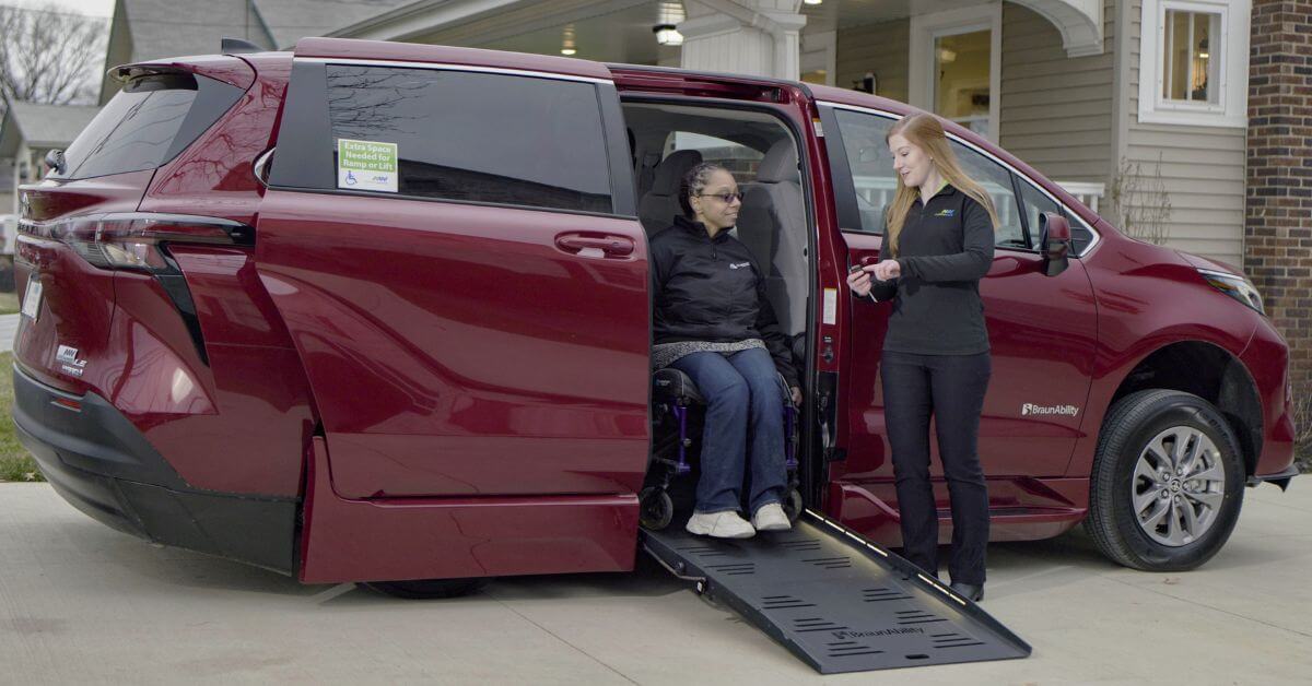 A woman in a wheelchair uses a ramp to exit a red minivan while another woman stands nearby holding keys. They are outside a house.