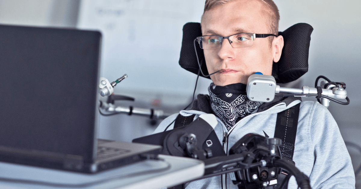 A man using a wheelchair and assistive technology with a mouth stick sits in front of a laptop, focusing on the screen.