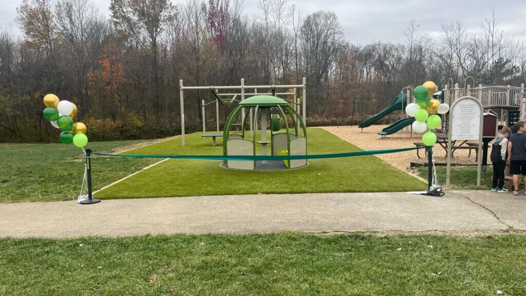 Un parque infantil con globos verdes y amarillos, que cuenta con una estructura para escalar y un tobogán, está acordonado con una cinta azul, con varias personas cerca.