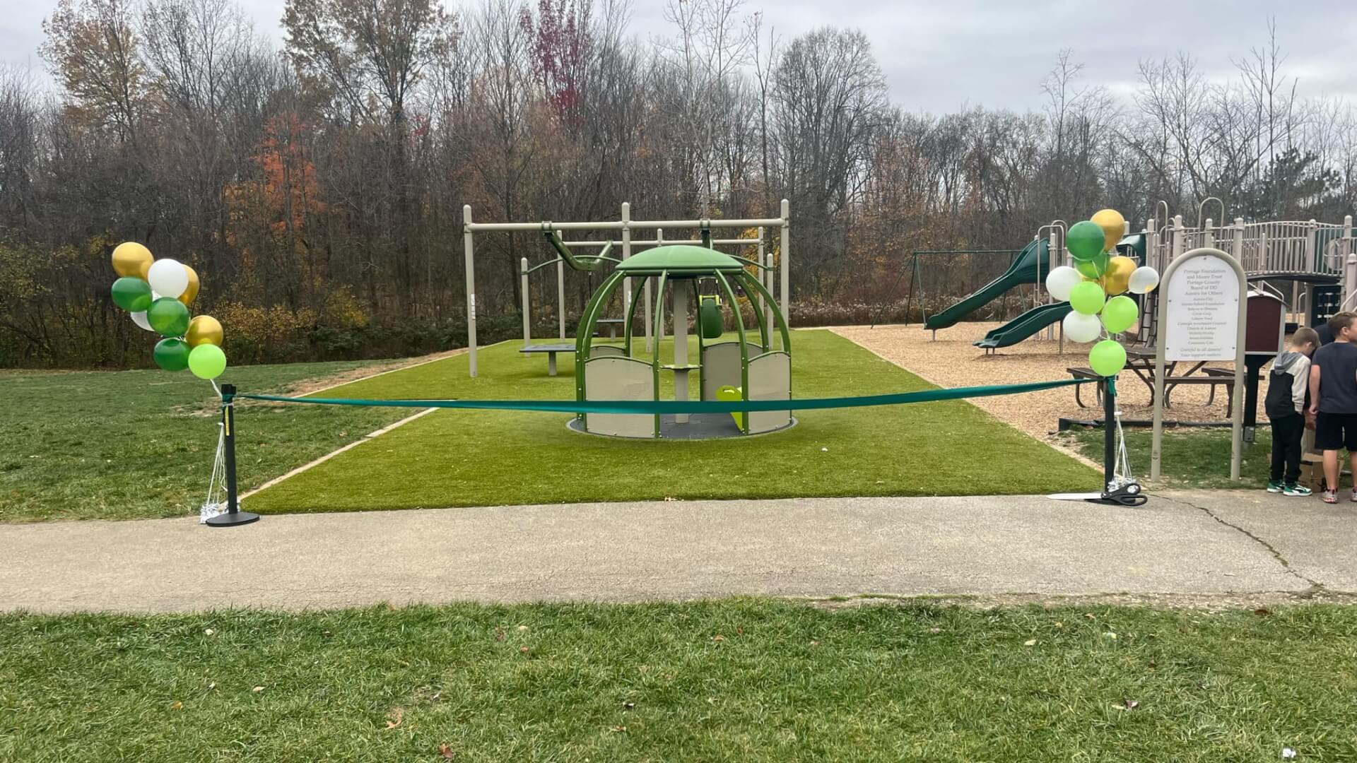 Un parque infantil con globos verdes y amarillos, que cuenta con una estructura para escalar y un tobogán, está acordonado con una cinta azul, con varias personas cerca.