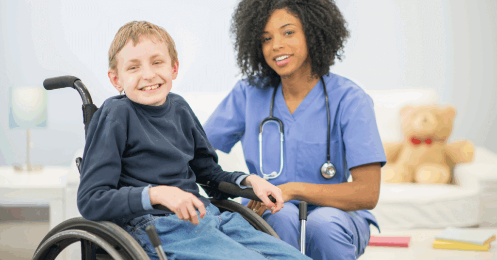 A smiling boy in a wheelchair sits next to a healthcare professional in blue scrubs, who has a stethoscope and is also smiling.