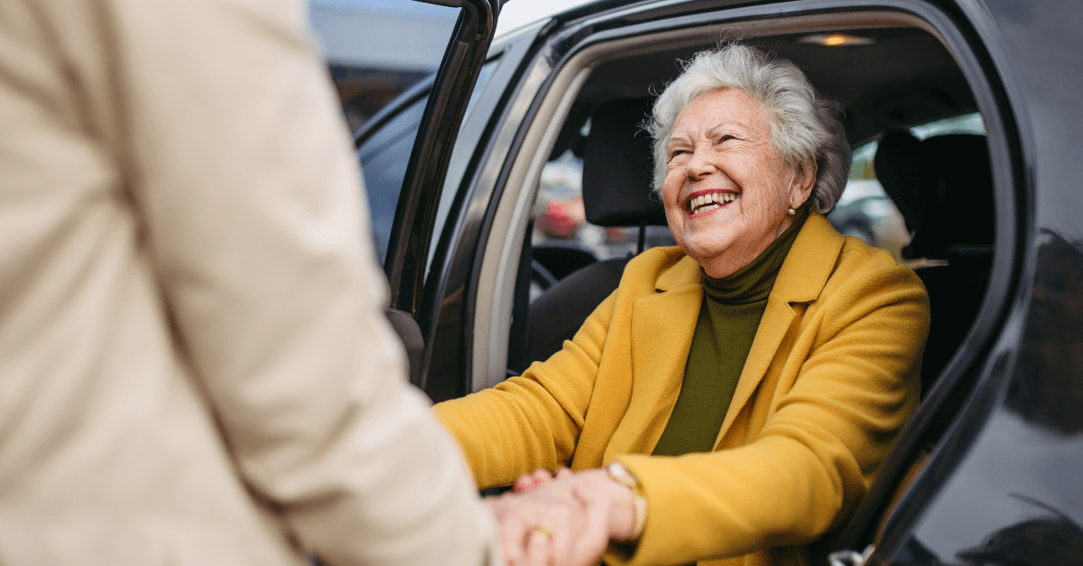 An older woman in a yellow coat smiles joyfully while exiting a car, holding hands with a person outside.