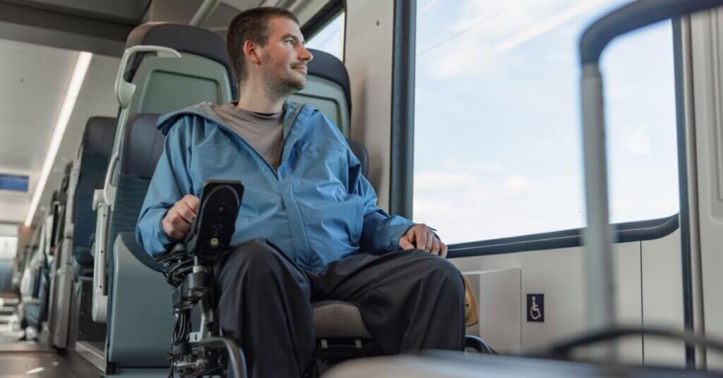 A man in a blue jacket sits in a wheelchair on a train, looking out the window. The train appears accessible and has modern seating.