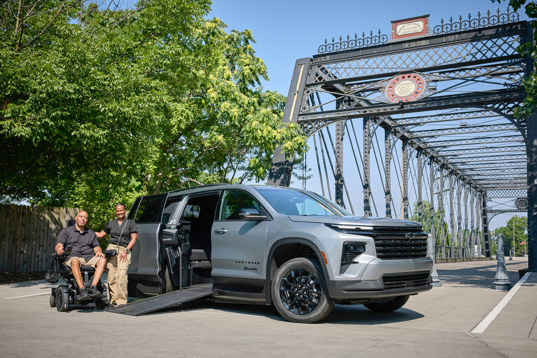 Two people, one in a wheelchair, are beside a gray wheelchair-accessible SUV with a ramp extended, parked near a historic iron bridge on a sunny day.