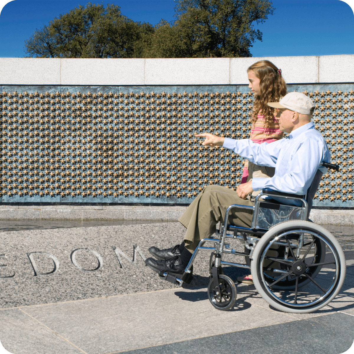 An older man in a wheelchair points while a young girl stands beside him at a memorial wall covered in gold stars, outdoors on a sunny day—an inspiring moment reminiscent of MobilityWorks promotions celebrating accessibility.