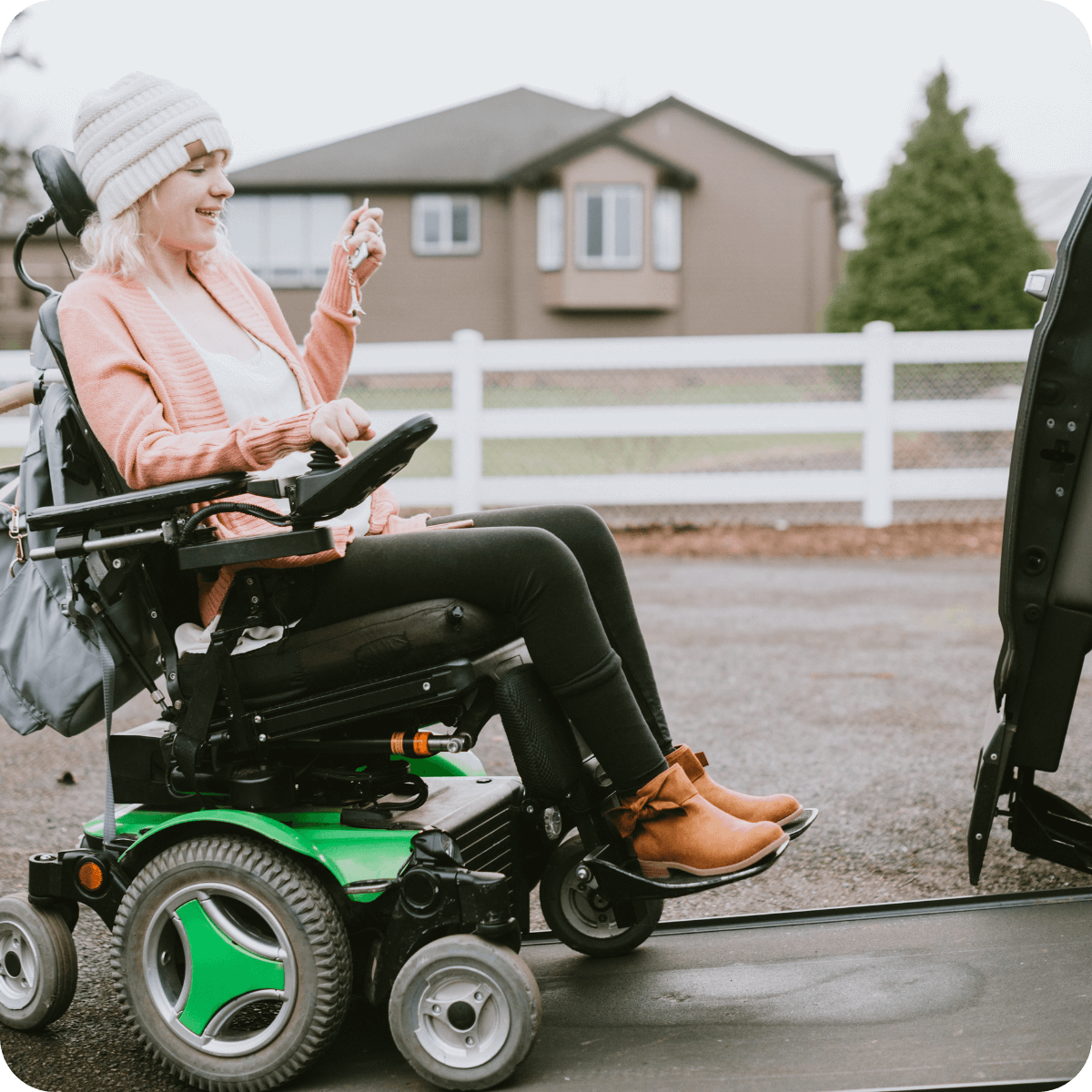 A woman in a motorized wheelchair holds car keys and smiles while using a wheelchair-accessible vehicle ramp outside a house.