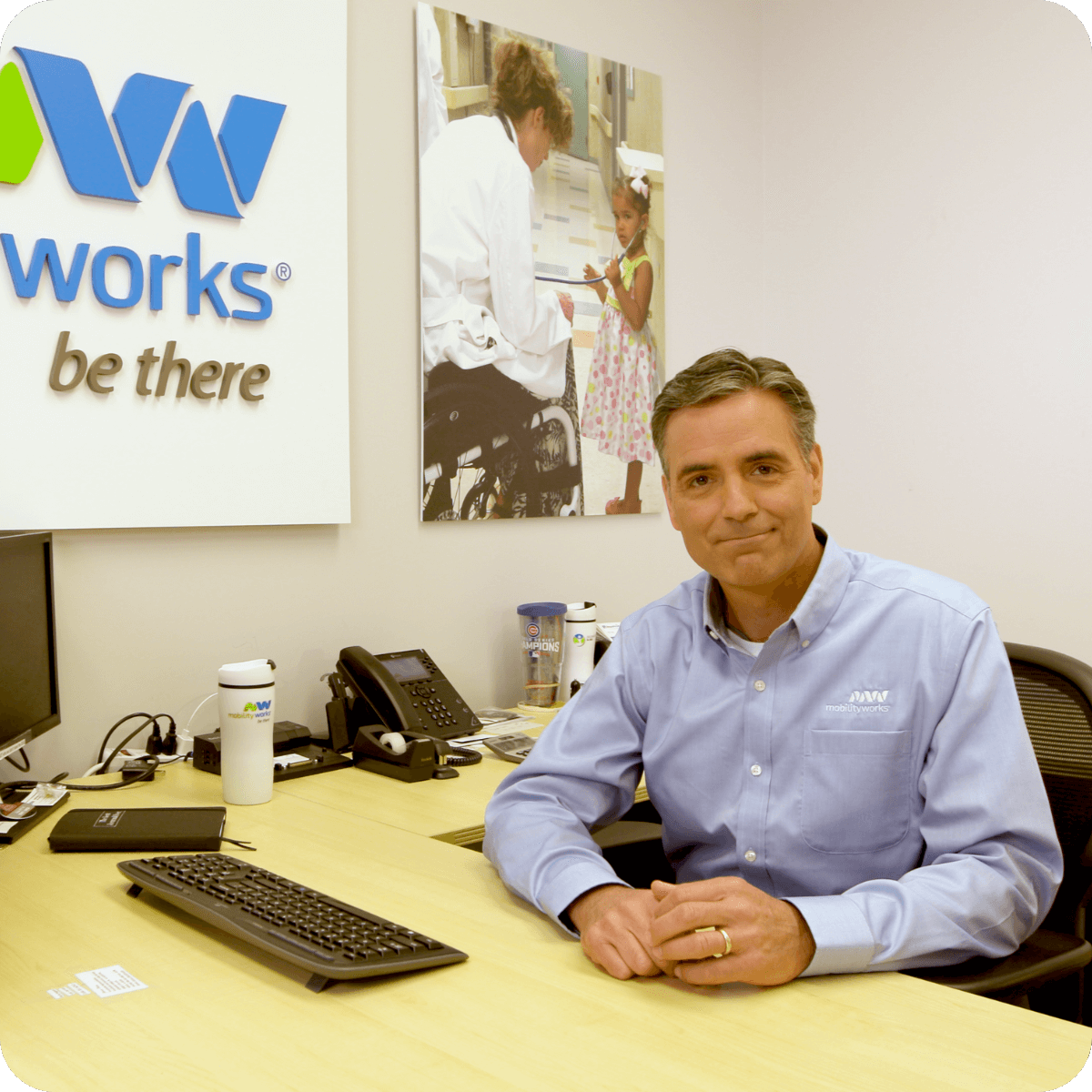 A man sits at an office desk, smiling at the camera. Behind him is a sign that reads wwworks® be there and a photo of a doctor with a child on the wall, possibly discussing the latest MobilityWorks promotions.