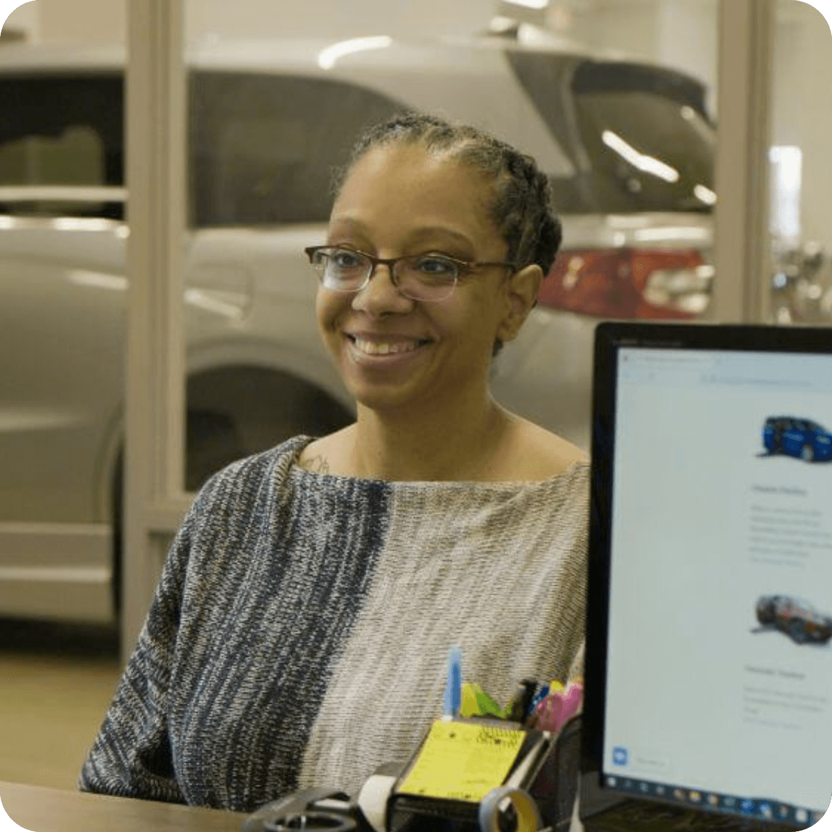 A woman with glasses and braided hair smiles while sitting at a desk in an office. A computer monitor and a car in the background hint at MobilityWorks promotions in progress.