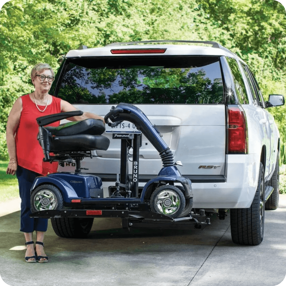 A woman stands beside a white SUV with a mobility scooter mounted on a platform lift attached to the vehicles rear.