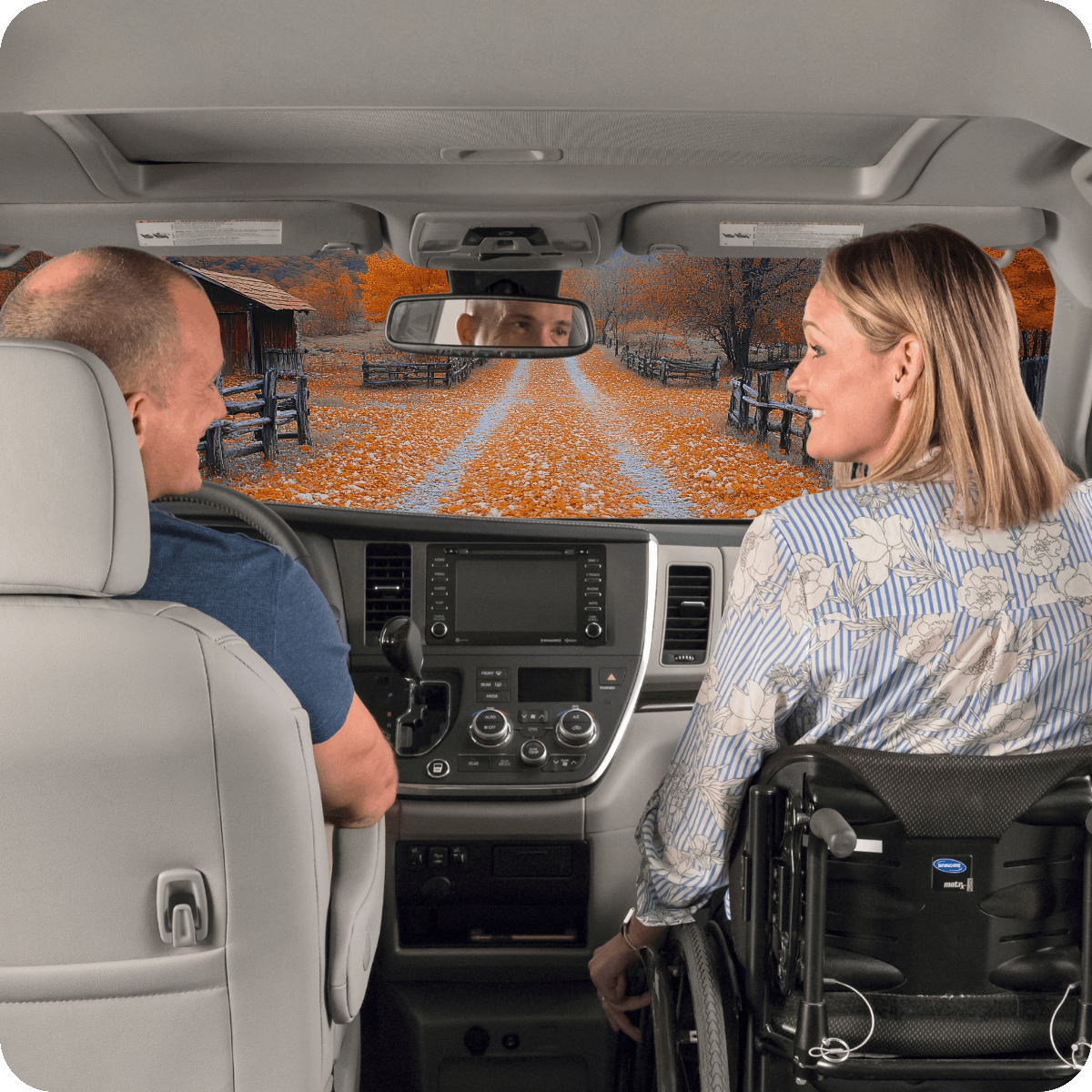 A man and a woman in a wheelchair are seated in the front seats of a van, looking at each other, with an autumn country road visible through the windshield.