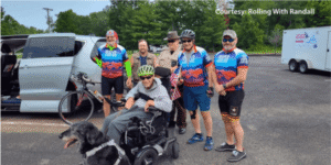 A group of cyclists, a man in a wheelchair with a dog, and two men in uniform pose in a parking lot near a van and trailer.