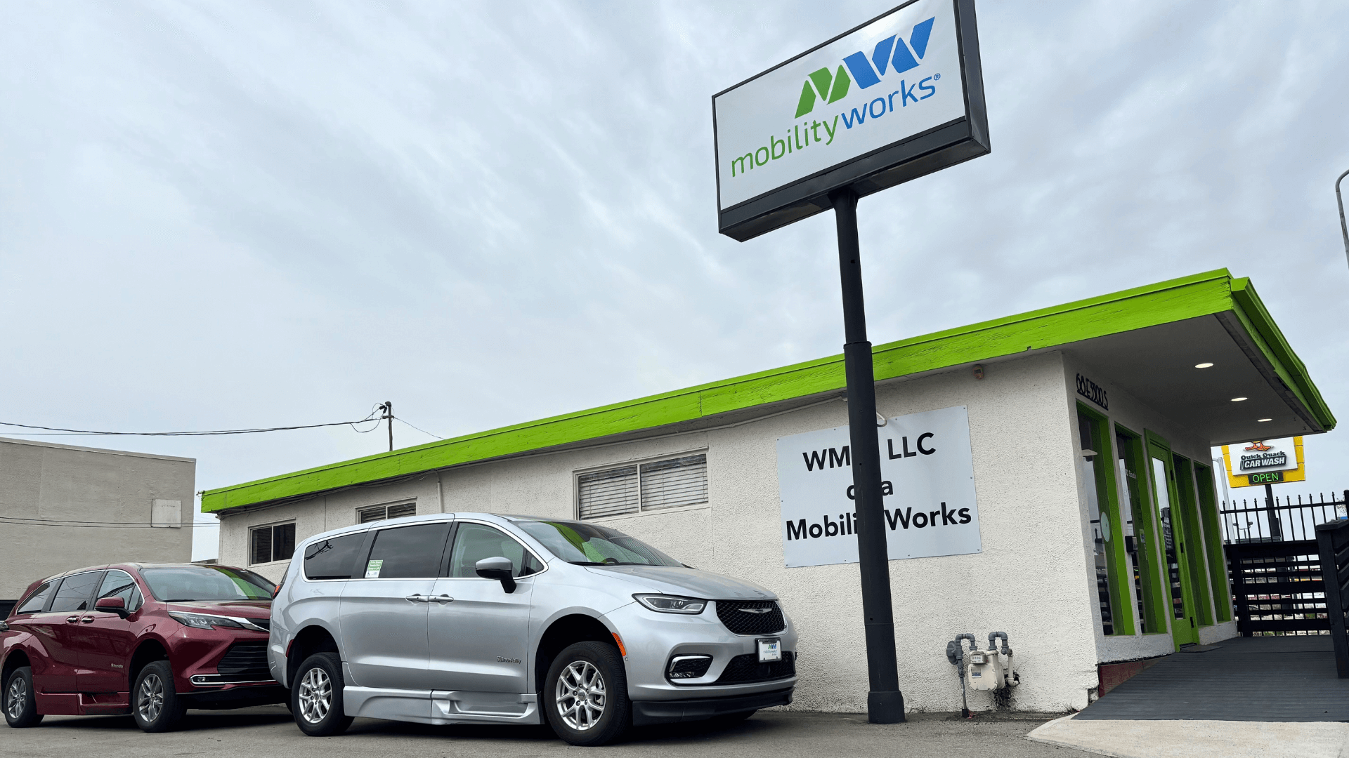 Exterior view of a MobilityWorks dealership showing parked minivans and a green-accented building under a cloudy sky.