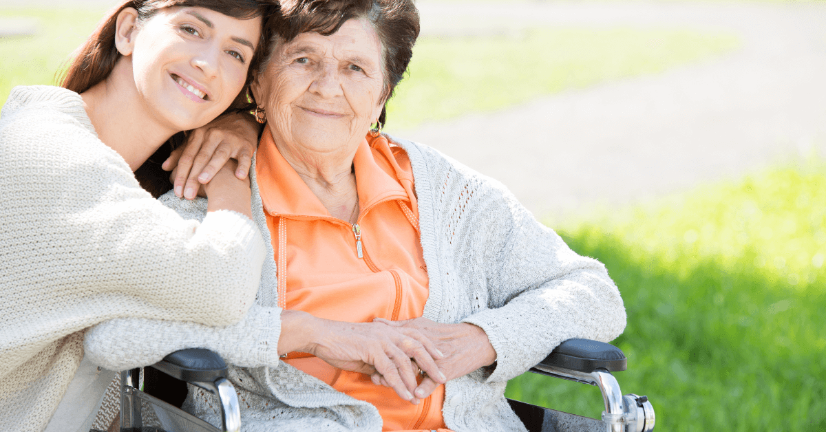 A young woman hugs an older woman sitting in a wheelchair outdoors on a sunny day. Both are wearing sweaters and smiling at the camera.