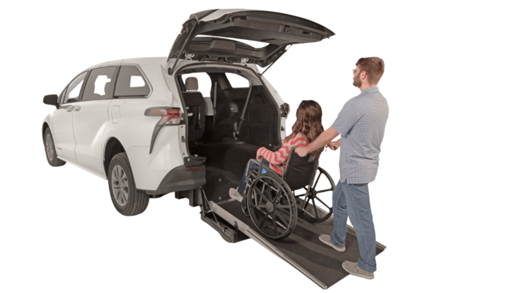 A man pushes a woman in a wheelchair up a rear-entry ramp into a white accessible van with an open back hatch.