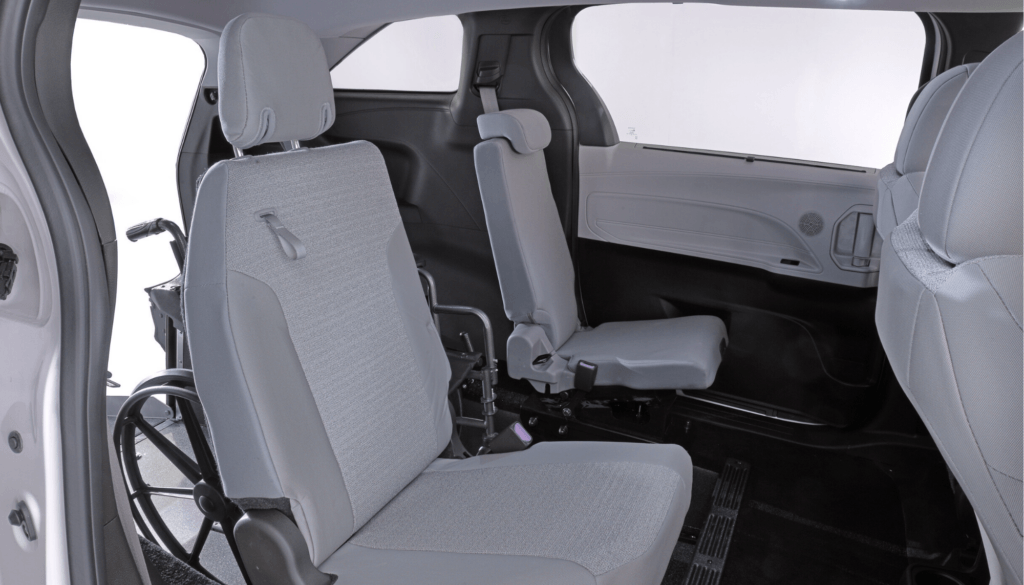 The image shows the interior of a vehicle with light gray seats, including a wheelchair secured in place behind the front passenger seat.