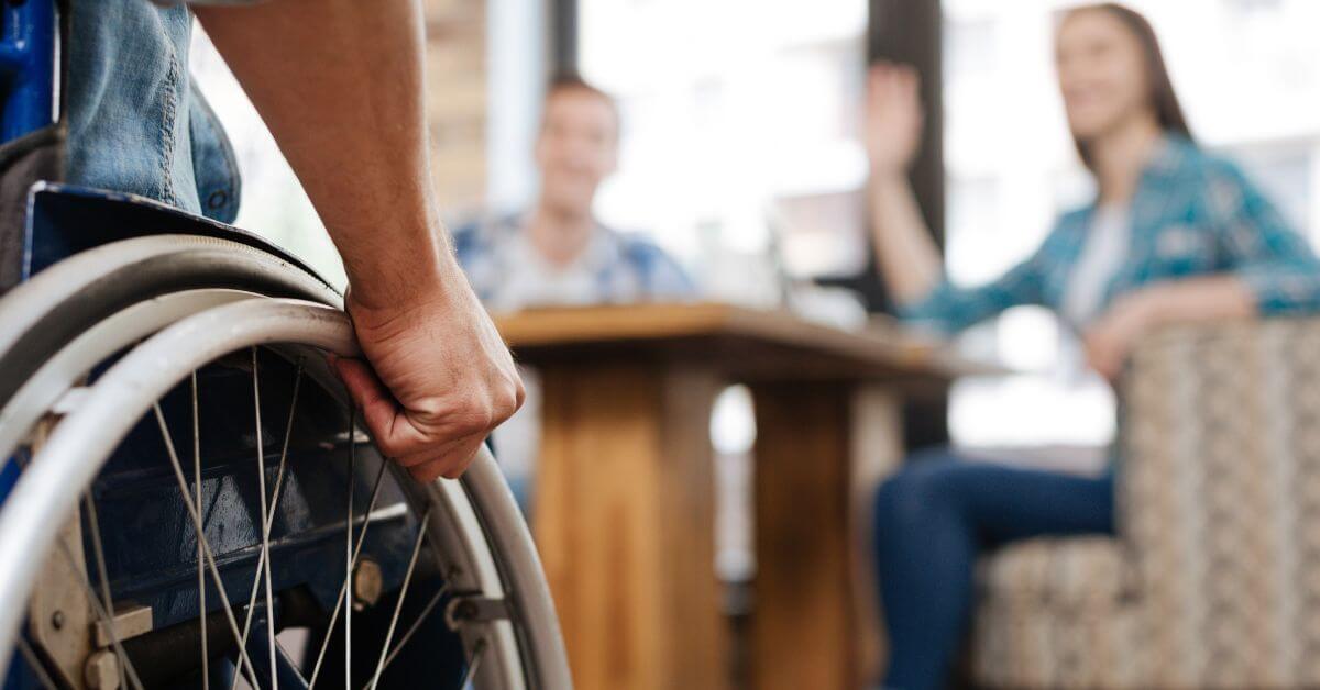 Person in a wheelchair approaches a table where two people are seated, one of whom is waving. The scene appears to be in a casual indoor setting.